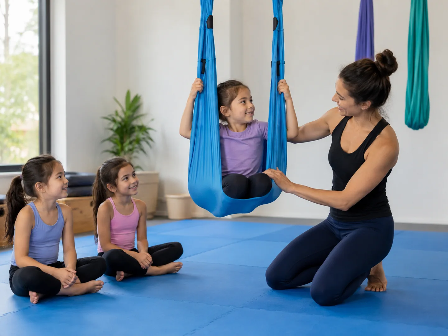An instructor guiding a child seated low in aerial fabric while classmates watch