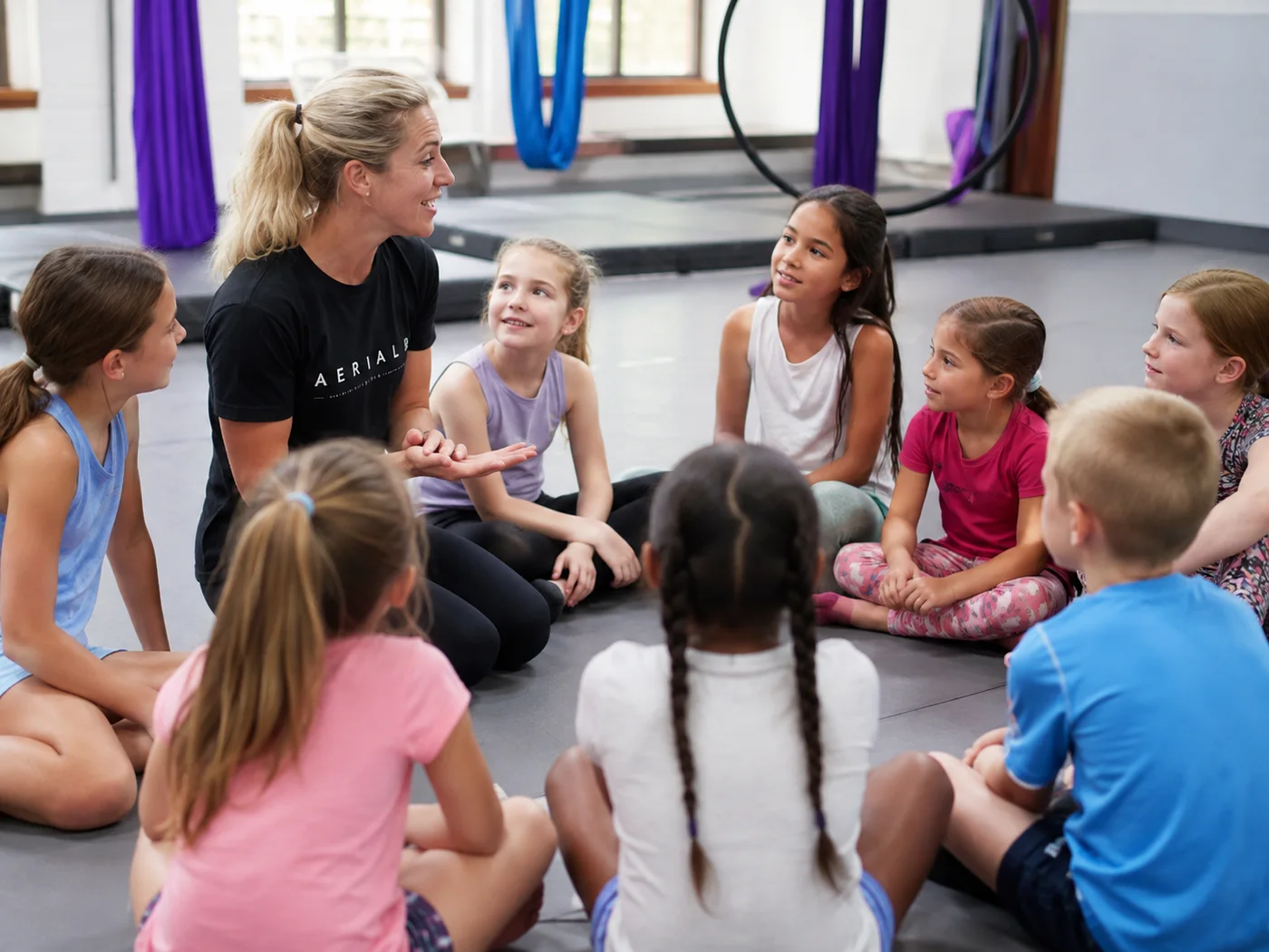 An aerial instructor talking with a seated group of children in a studio